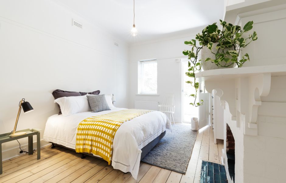 White room with brown wooden flooring. There is a bed with a white bedspread and grey rug beneath. A single bulb hangs over the room