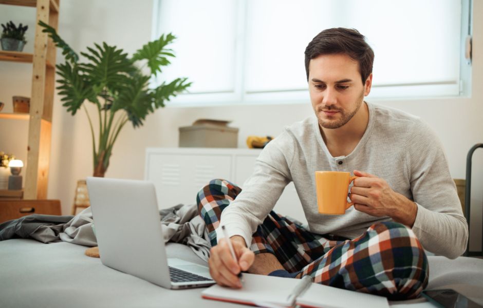 A man sits on a his bed in his pyjamas. He is using a laptop and holding a yellow coffee cup. 