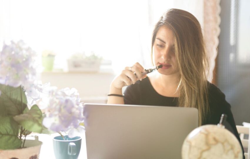 A woman sits at a laptop - she has a vape in her mouth.