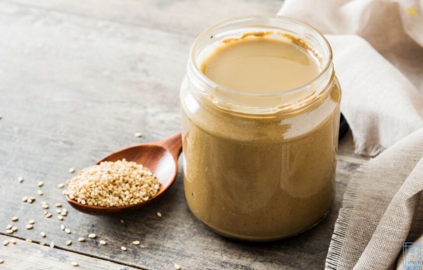 A jar of golden coloured tahini paste with a spoon filled with sesame seeds next to it