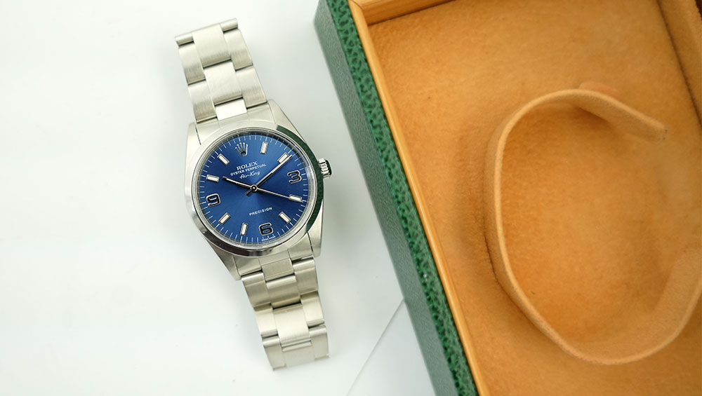 A silver Rolex watch with a blue face placed next to an open green watch box, which has a orange lining