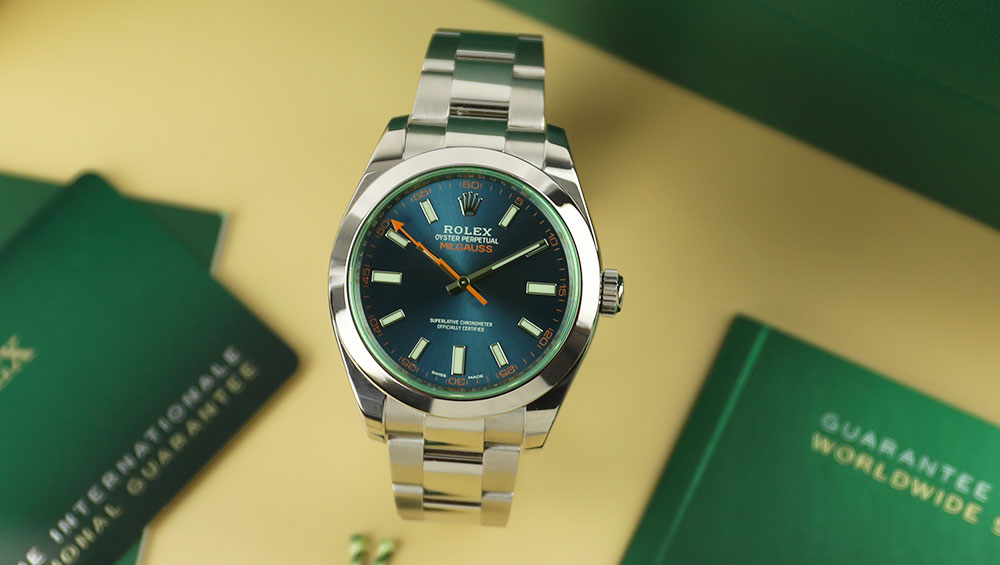 A silver Rolex watch with a blue/green face and light green details. One of the clock hand is orange. There are green pamphlets in the background.