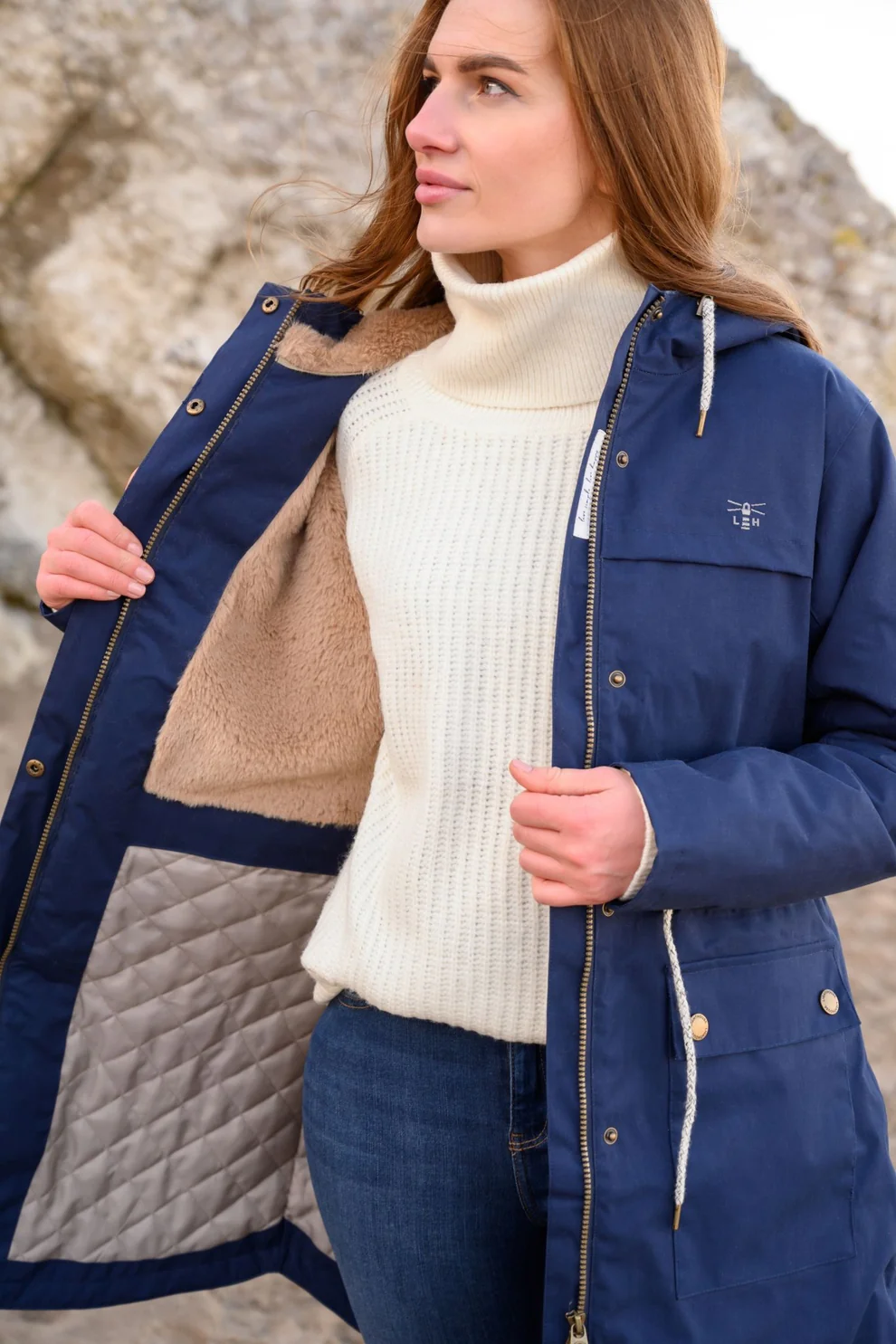 A woman is wearing a blue coat and white polo neck jumper. She is holding the coat open to show the fleecy inside layer