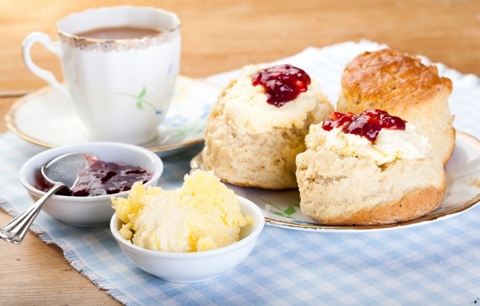 A classic cream tea; a white cup a saucer filled with tea, scones topped with cream and jam, one uncut scone and pots containing cream and jam, all placed on a blue and white checked cloth