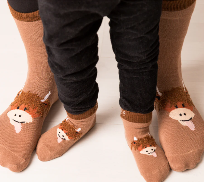 Two pairs of feet - one big and small wearing brown socks which feature a cartoon highland cow