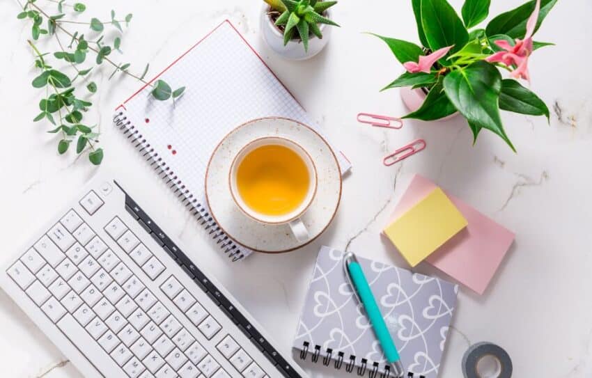 A flat lay of a computer keyboard, post-it notes, note books, pink paper clips and a cup containing a yellow drink. There are also three plants and some tap.