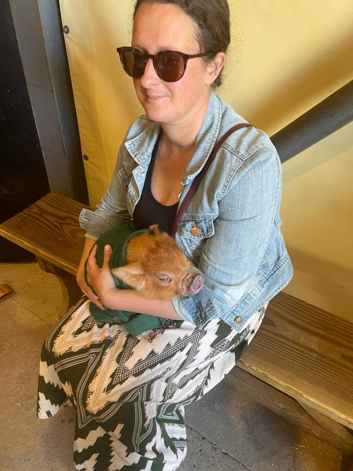Fee (blog owner) cuddling a piglet wrapped in a green blanket. She is wearing a patterned dress a denim jacket and sun glasses