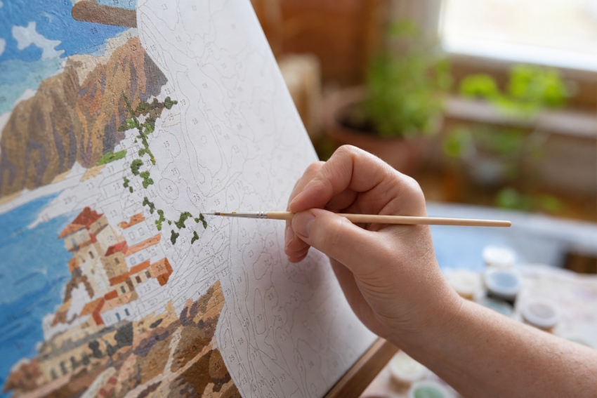A close-up view of someone painting on a highly detailed numbered canvas with a wooden brush